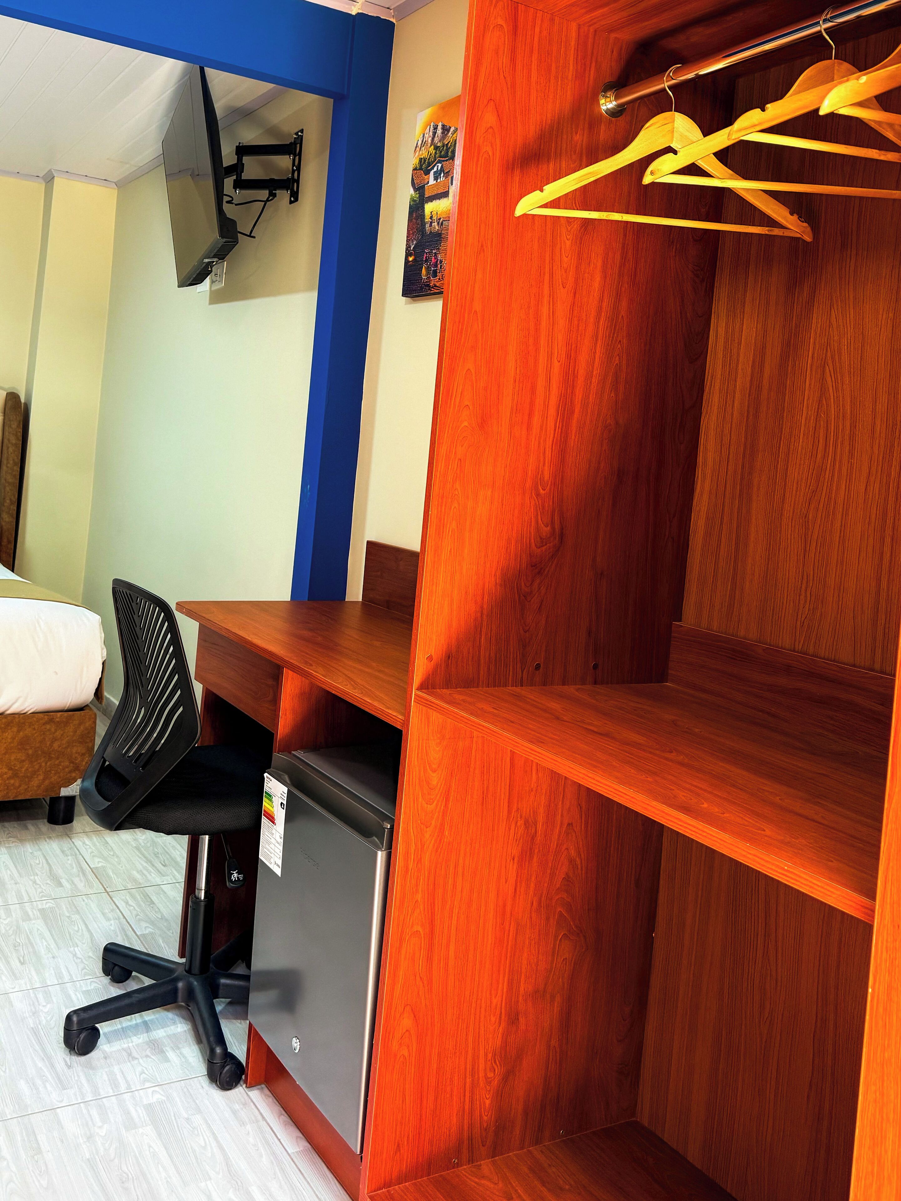 A room with a wooden desk, a black office chair, a bed, and a wall-mounted TV.