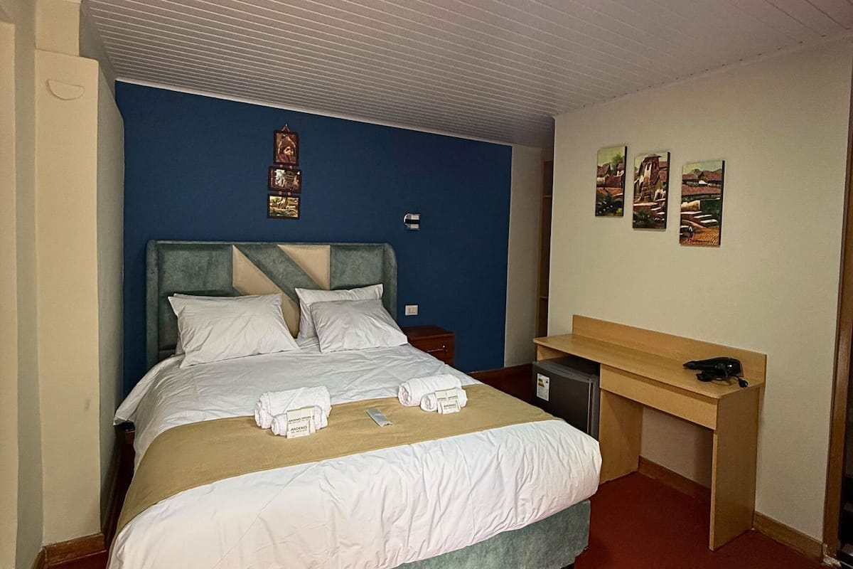 A neatly made bed with white linens and a grey headboard, a small desk with a chair, and a red carpeted floor.