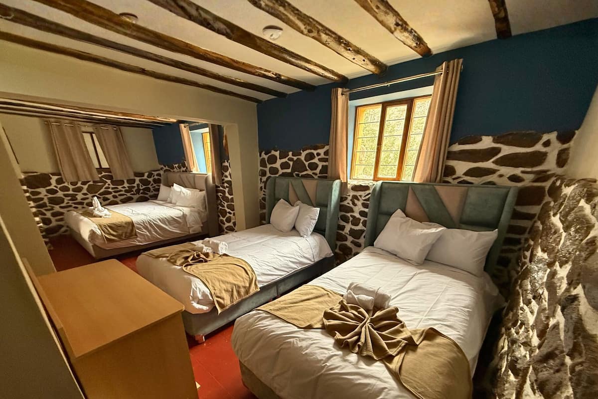 A room with two beds, stone walls, a wooden ceiling, and a window with curtains.