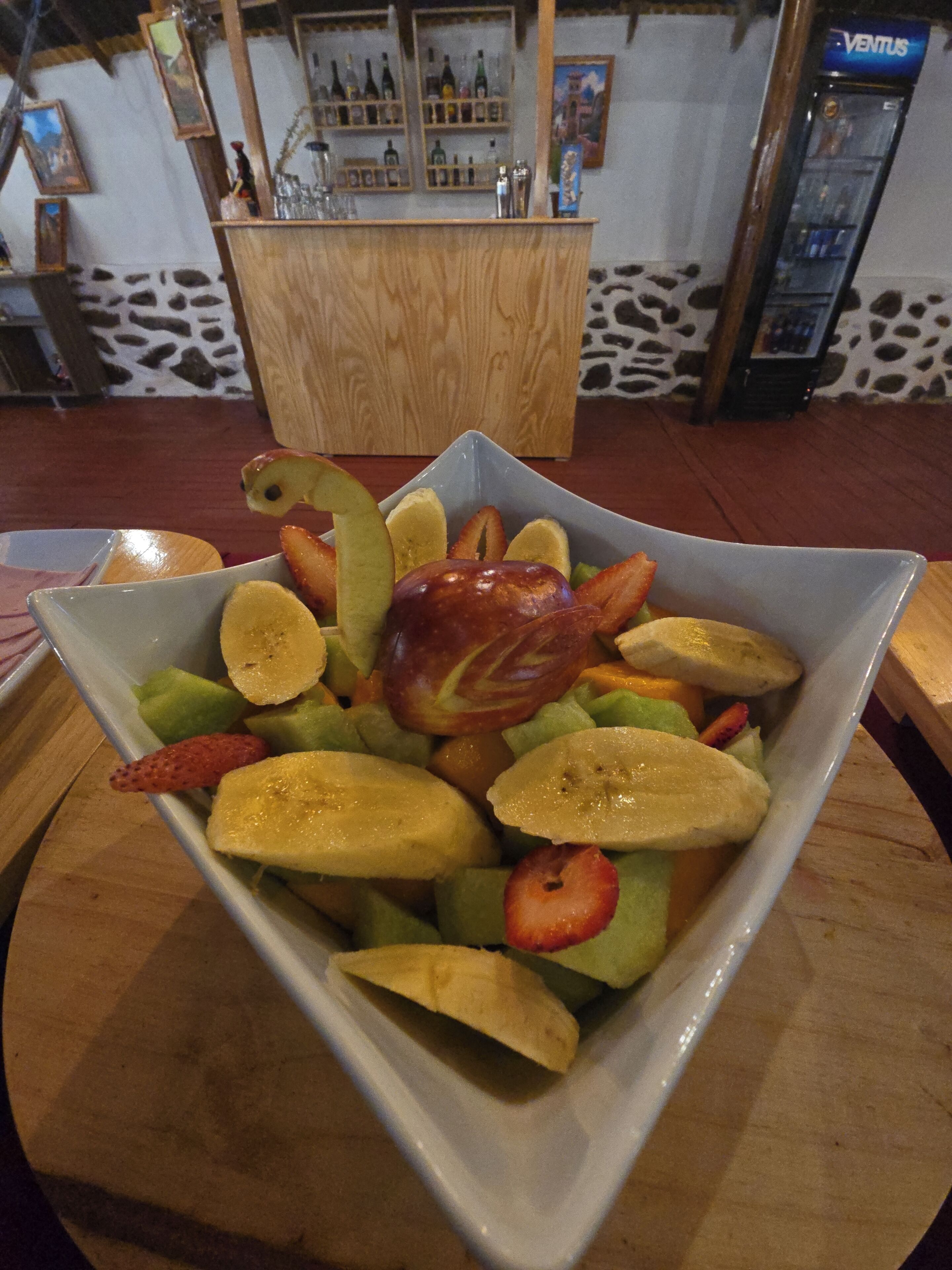 A square bowl filled with sliced fruits and a decorative apple on a wooden table.