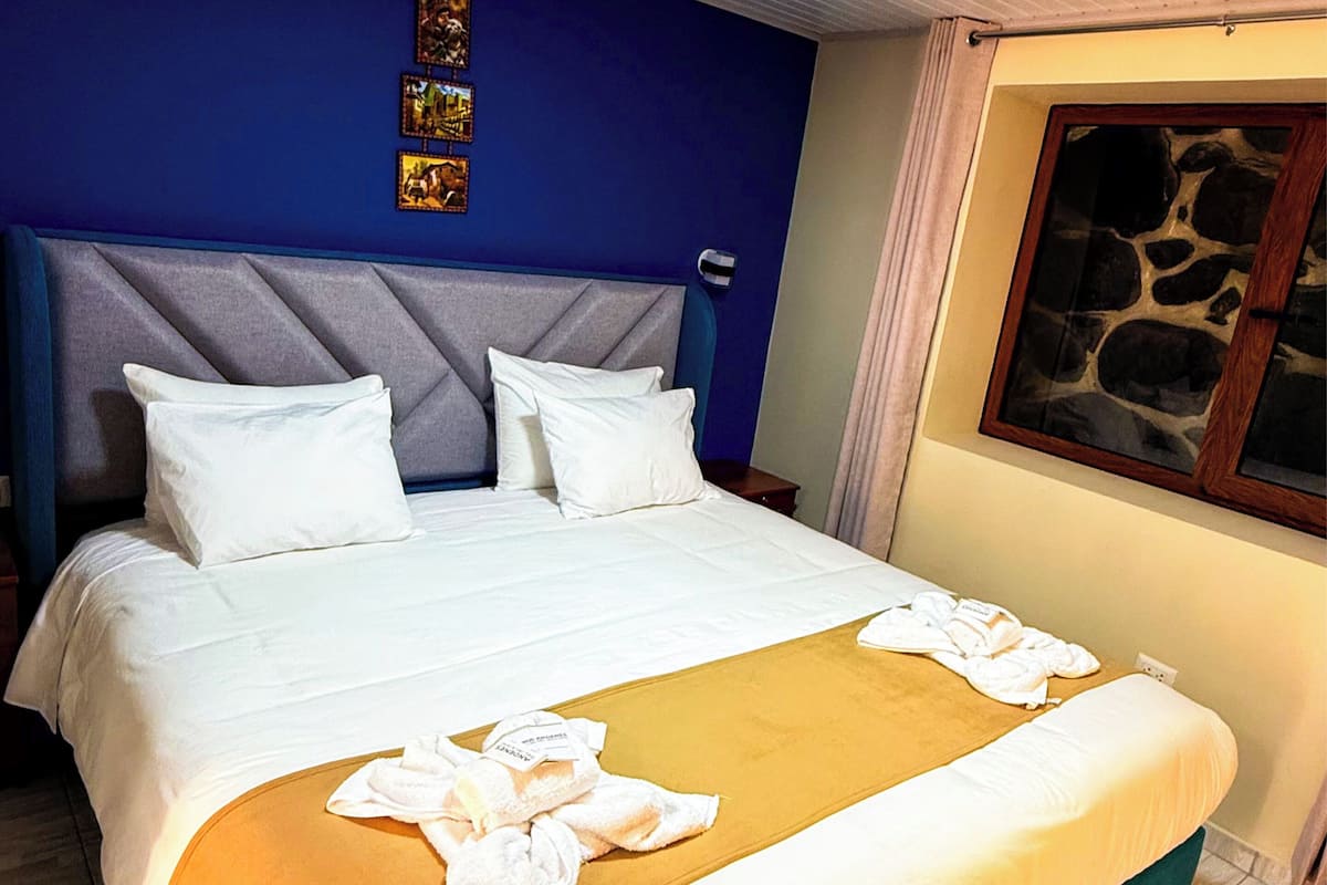 A neatly made bed with white linens and a yellow throw blanket, a blue headboard, and a stone-patterned window.