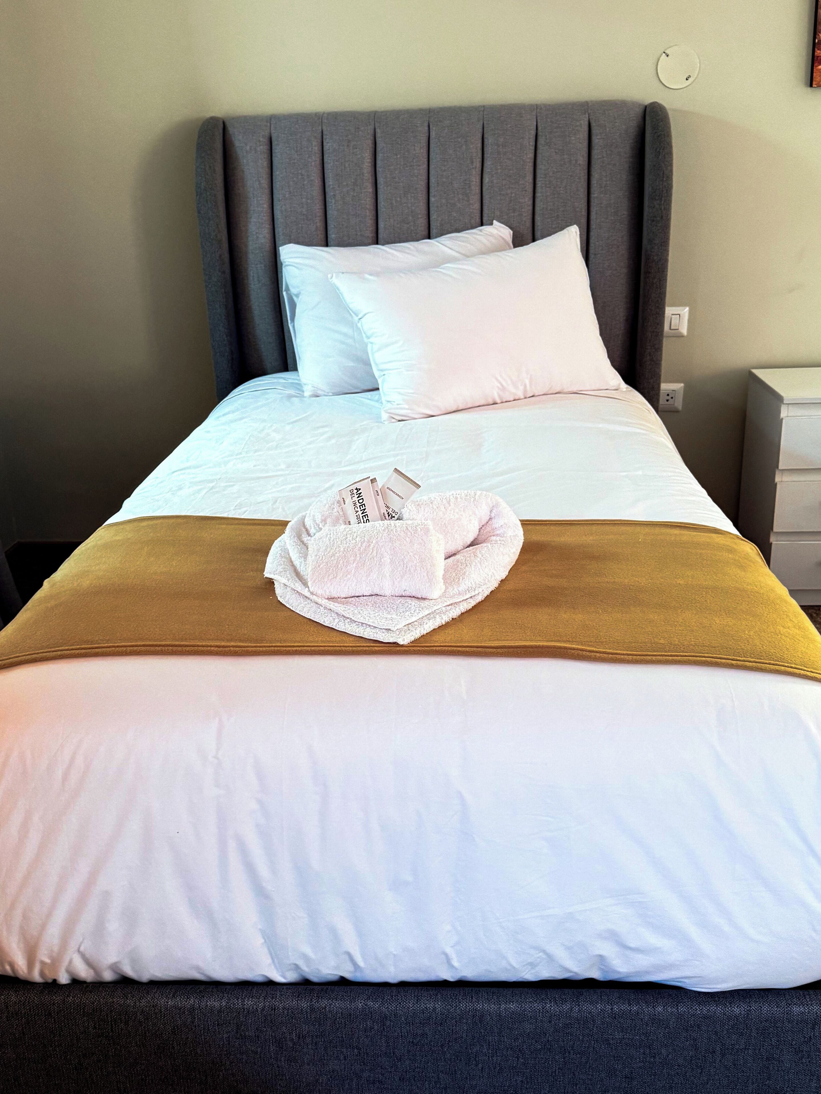 A neatly made bed with white linens, a brown throw blanket, and a white towel with text on it.