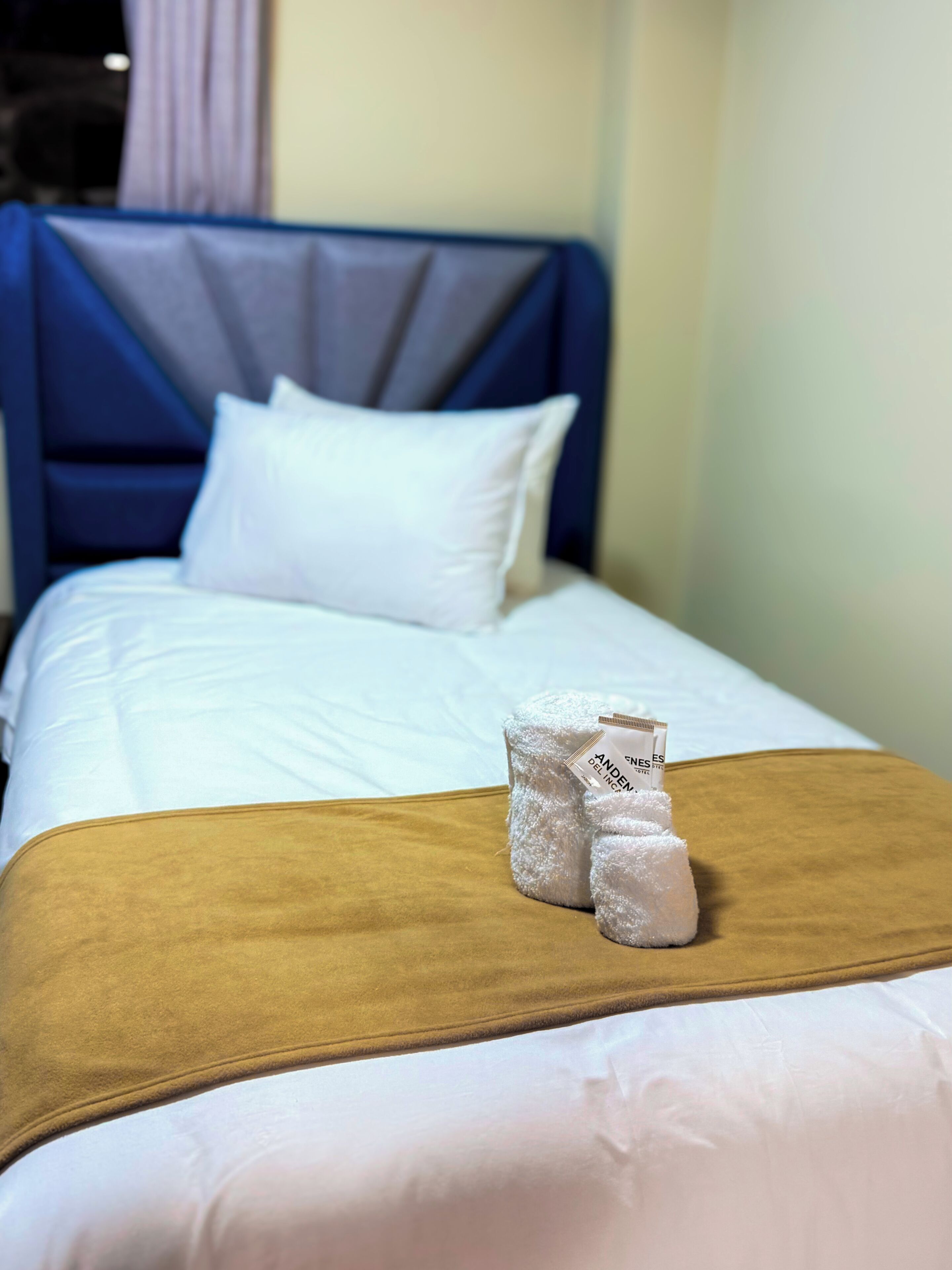 Una habitación de hotel con una cama, ropa de cama blanca, un cabecero azul y una manta marrón.