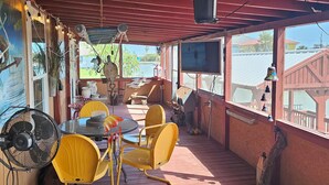 A covered outdoor seating area with yellow chairs and a table, a fan, and a TV screen.