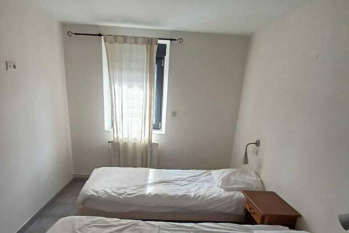 A small, clean room with two single beds, a window with curtains, a bedside table, and a wall-mounted light fixture.