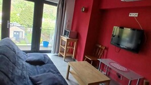 A living room with a sofa, a wooden table, a TV mounted on the wall, and a window with curtains. - Presqu'île de Chooz (Chooz)