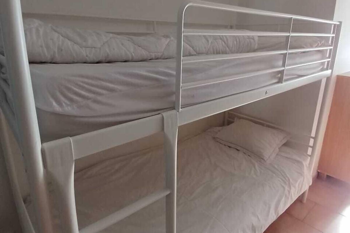 A bunk bed room with a white metal frame, white bedding, and a small wall-mounted light fixture.