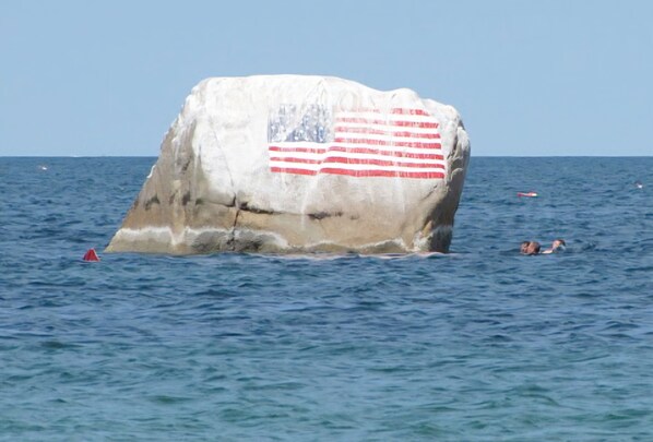 Una gran roca con una bandera estadounidense pintada, rodeada de agua y algunas personas nadando.