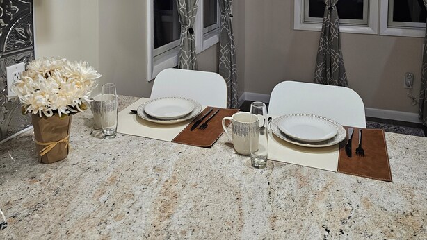 Een keuken met granieten aanrechtbladen, witte stoelen, een eettafel voor twee personen en een bloevas.