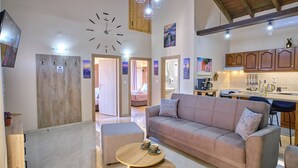 Un saló modern amb un sofà, una otomana i una taula de centre. Hi ha una zona de cuina amb armaris de fusta i una taula de dinar. Es veuen un rellotge i llums penjants al sostre.