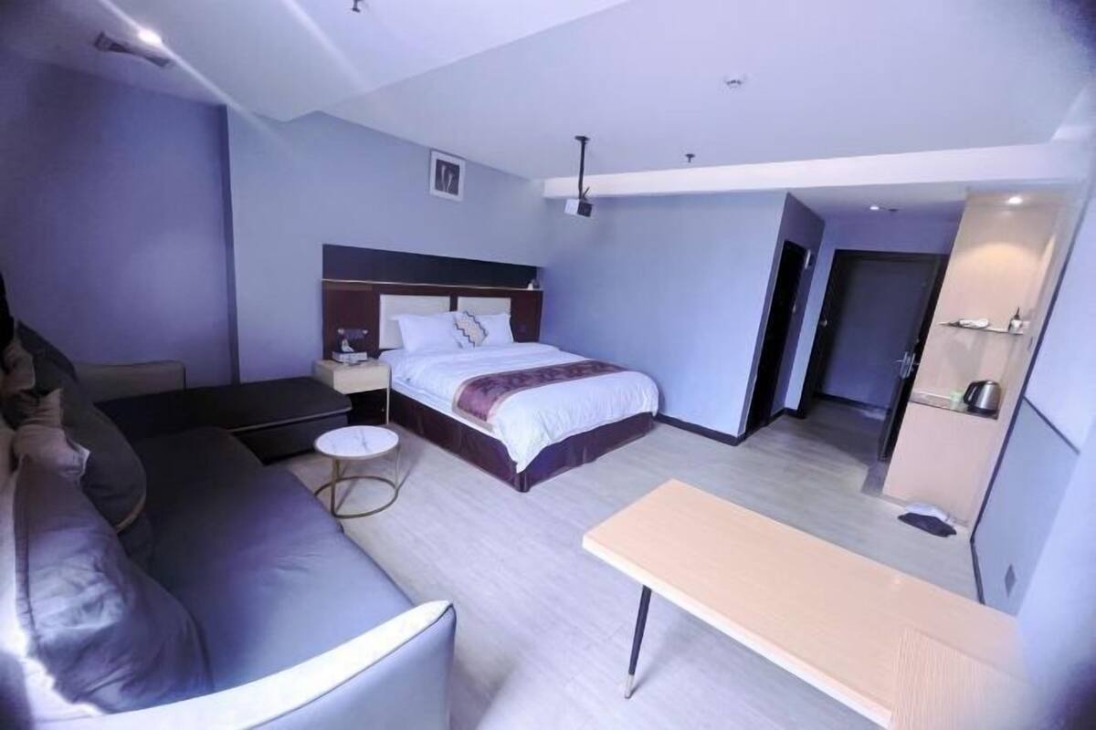 A modern hotel room with a bed, a sofa, a small round table, and a kitchenette area.