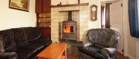 A living room with a wood-burning stove, a dark patterned sofa, a wooden coffee table, and a patterned armchair.