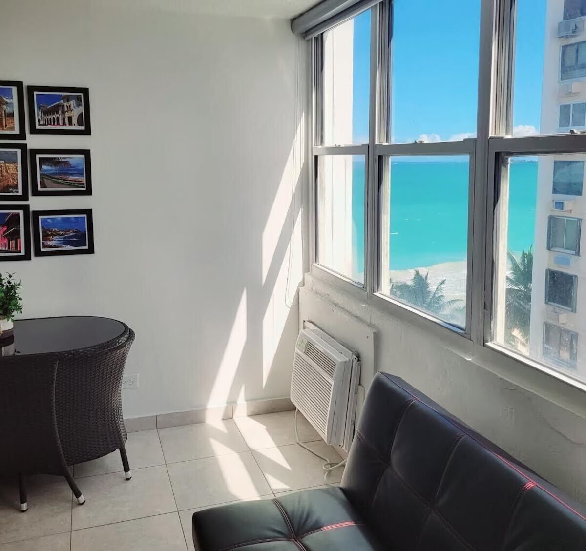 A room with a view of the sea, a dining table, a sofa, and several framed pictures on the wall.