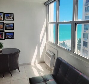 A room with a view of the sea, a dining table, a sofa, and several framed pictures on the wall.
