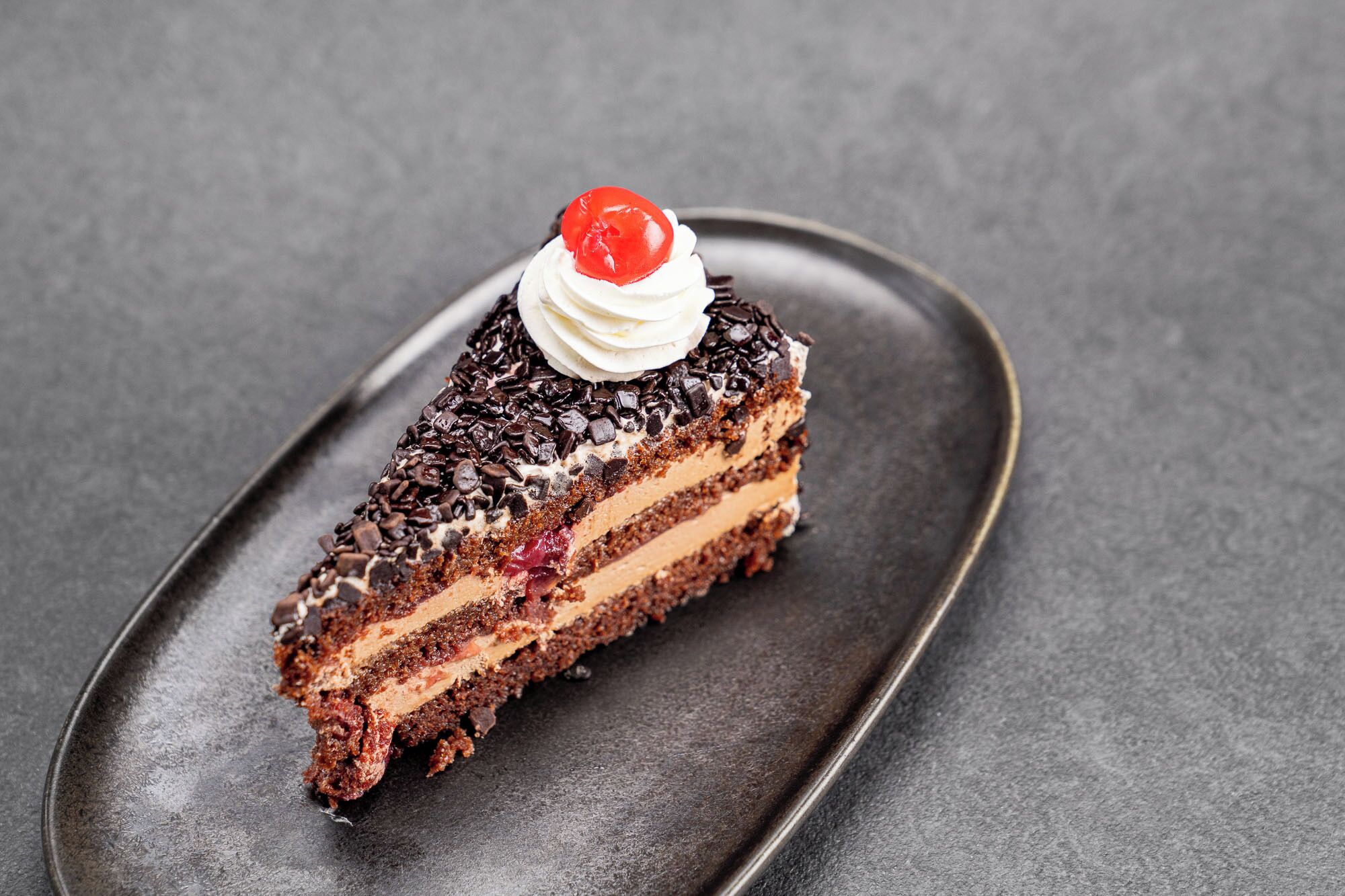 Una rebanada de pastel de chocolate en capas con crema batida y una cereza encima, sobre un plato oscuro.