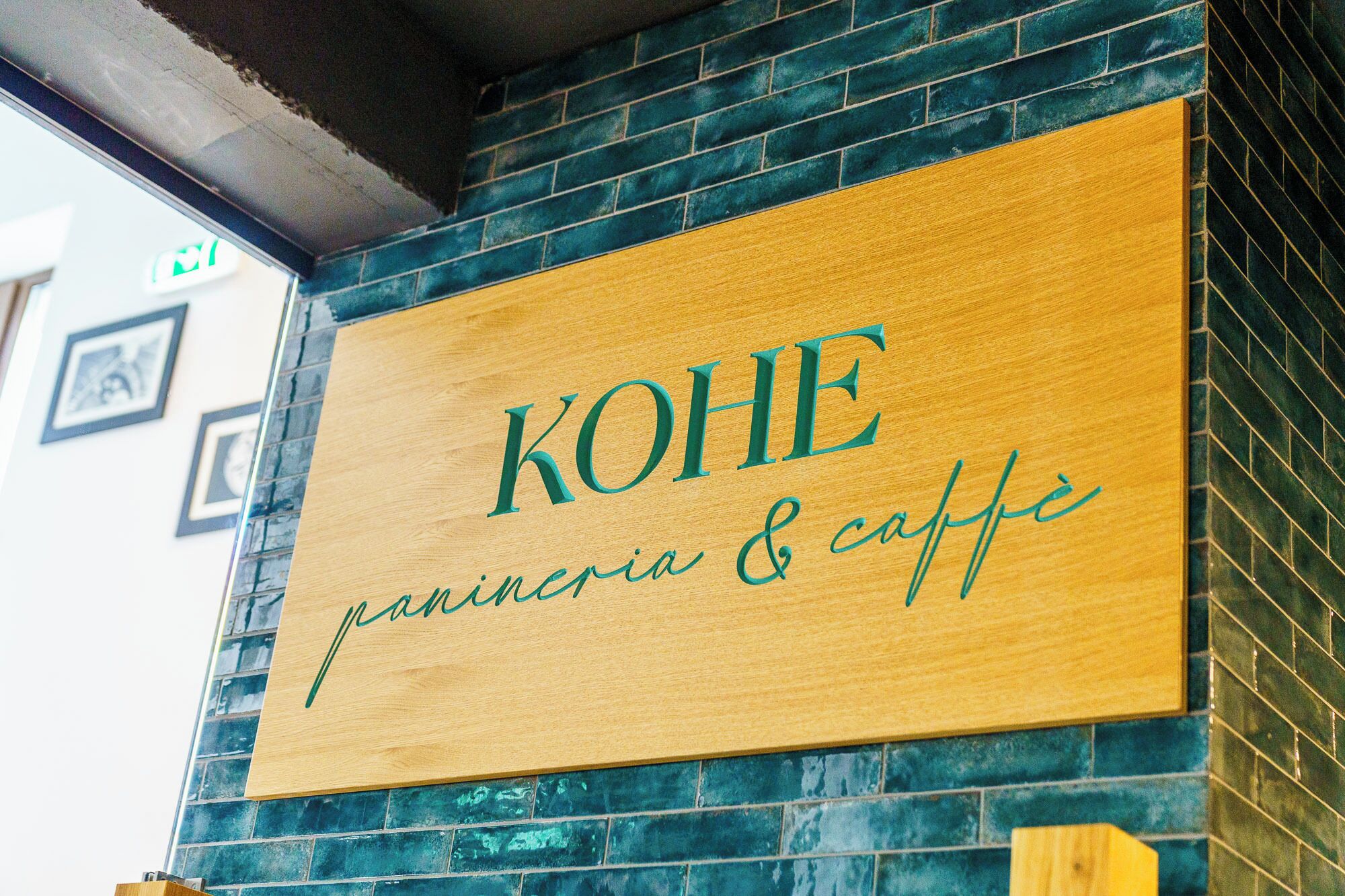 Un letrero de madera con la inscripción "KOHE panineria & caffè", colocado sobre una pared con azulejos azules.