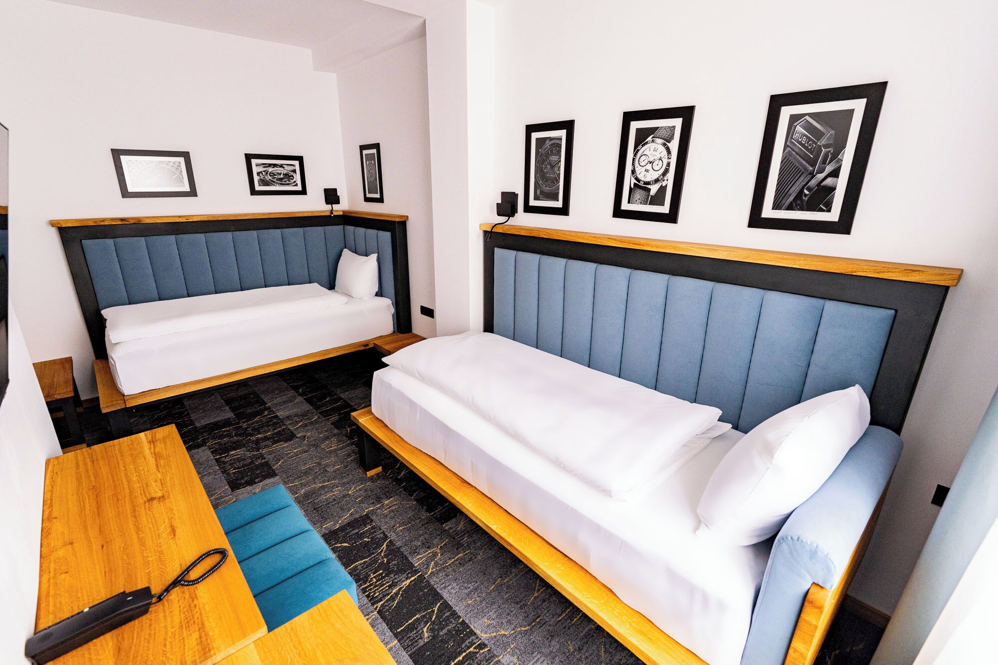 Habitación de hotel con dos camas individuales, una mesa de madera y un banco azul. En la pared hay fotografías enmarcadas.