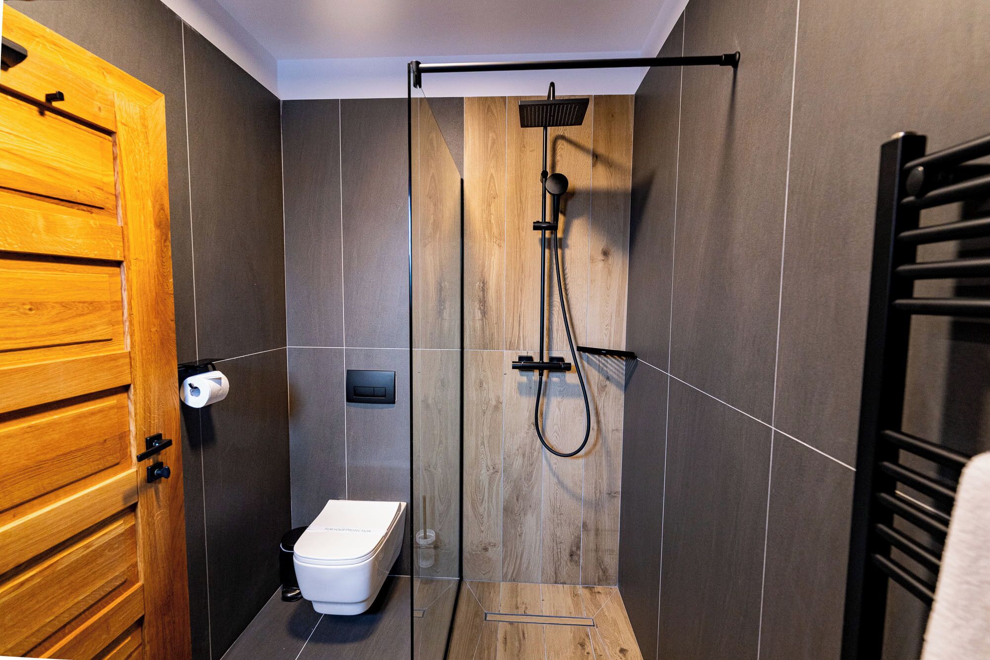 Un baño moderno con puerta de madera, inodoro, ducha con cabezal de lluvia y un perchero para toallas.