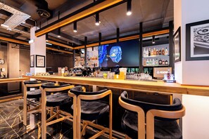 A bar area with wooden bar stools, a counter with a digital screen displaying a watch, and shelves with various bottles. - HOW Boutique and Hotel (Cluj-Napoca)
