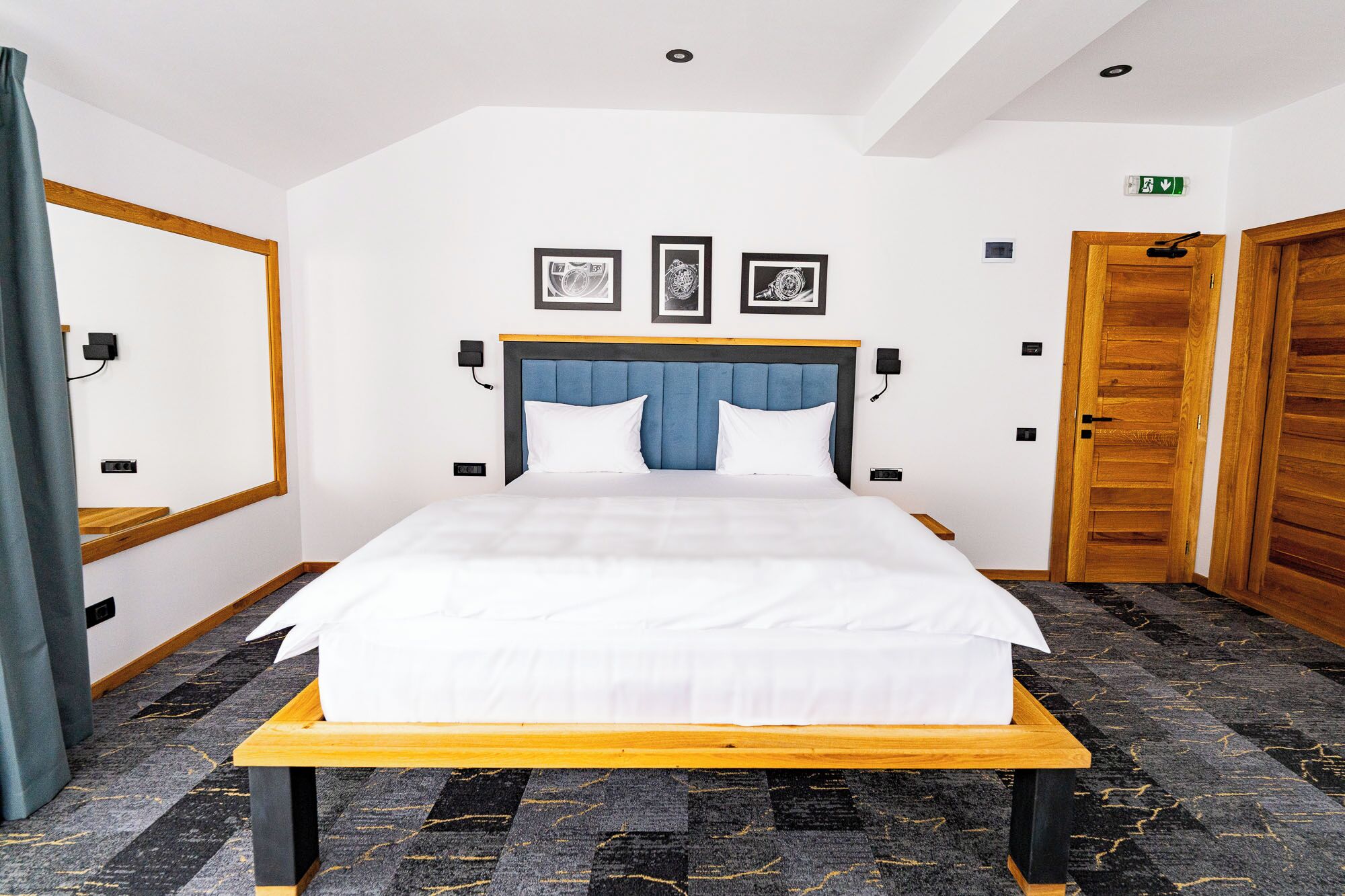 A hotel room with a bed, a wooden headboard, a blue accent, framed pictures on the wall, and a wooden door.