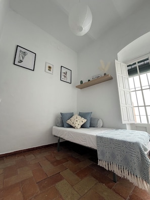 Una habitación con cama, ventana con contraventanas, lámpara en el techo e imágenes colgadas en la pared.