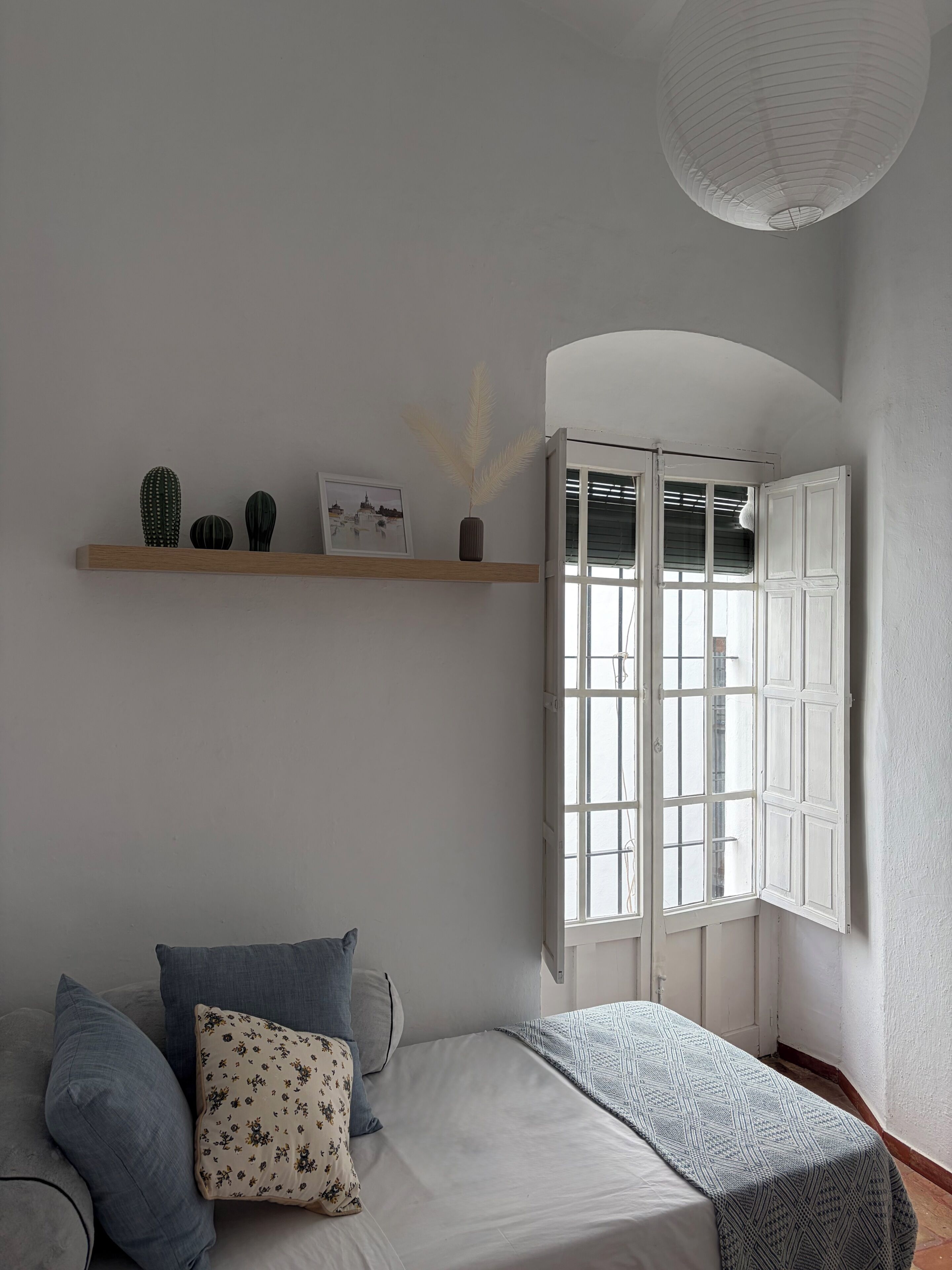 Un dormitorio con cama, almohadas, una ventana con contraventanas abiertas, una estantería con adornos y una lámpara colgante.