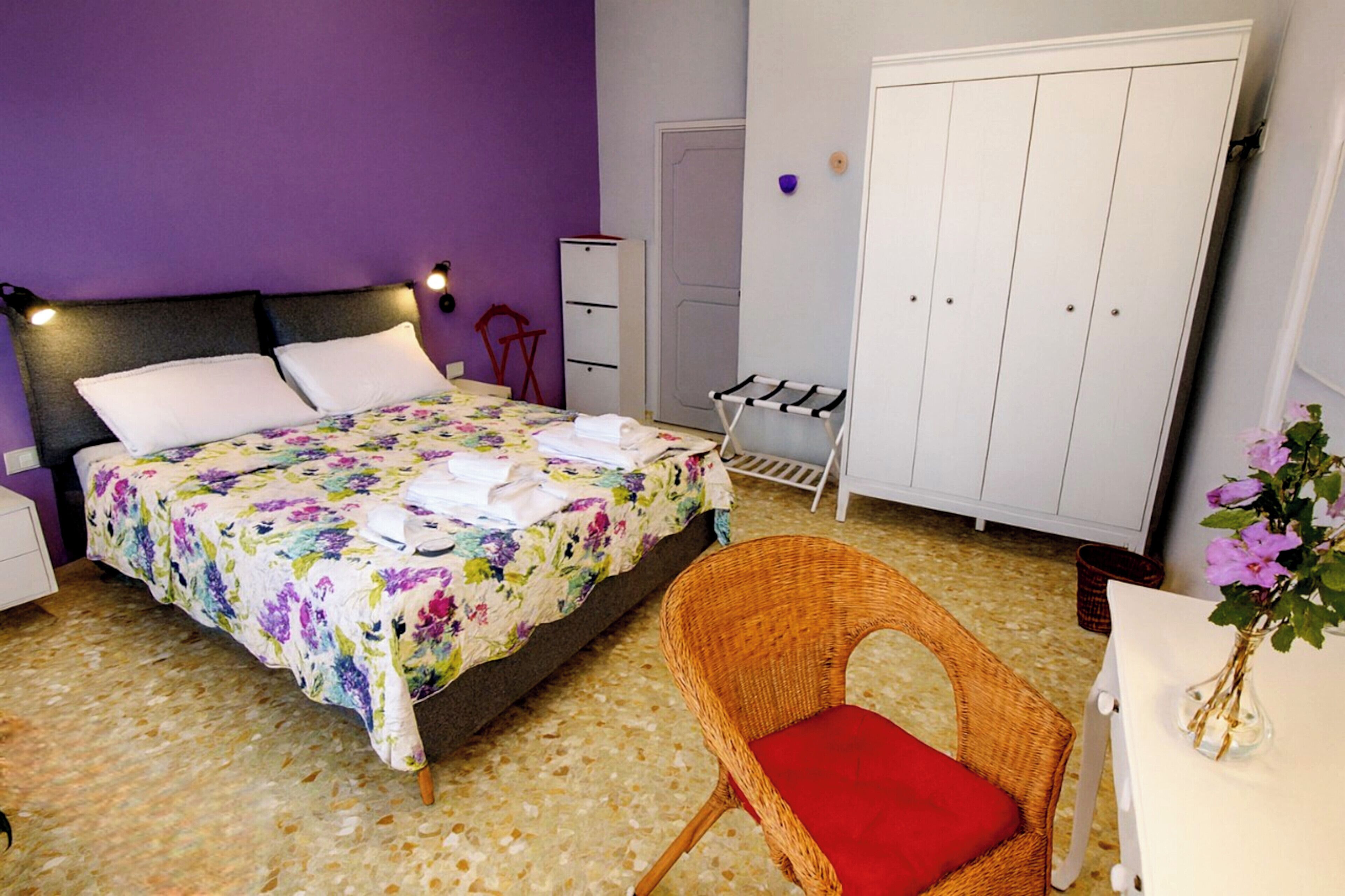 A bedroom with a bed, a wicker chair, a wardrobe, and a small table with a vase.