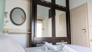 Quarto de hotel com um espelho grande, uma cama com toalhas brancas bem dobradas, um guarda-roupa de madeira e um pequeno armário com produtos de higiene pessoal.
