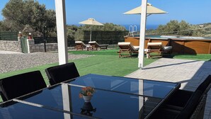 Uma mesa com tampo de vidro e cadeiras de vime, vista para uma piscina e guarda-sóis, e um muro de pedra ao fundo.