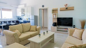 Uma sala de estar moderna com um sofá bege, uma mesa de centro e uma televisão. Dá para ver uma cozinha ao fundo, com armários brancos e um espaço para refeições.