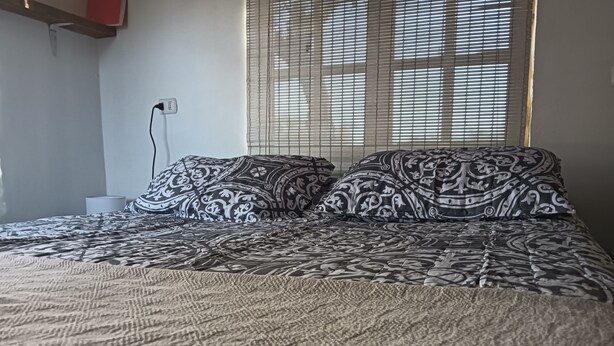 A bed with patterned bedding, a power outlet, and a window with blinds.