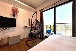 Habitación de hotel con cama, televisión montada en la pared, ventilador de pie, una silla y balcón con vistas.