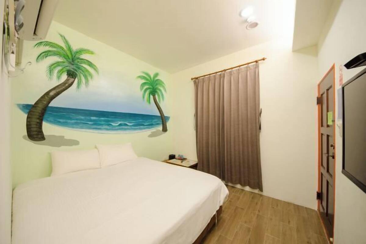 Una camera da letto con un letto, un comodino, una TV, una tenda e un murale raffigurante una spiaggia.