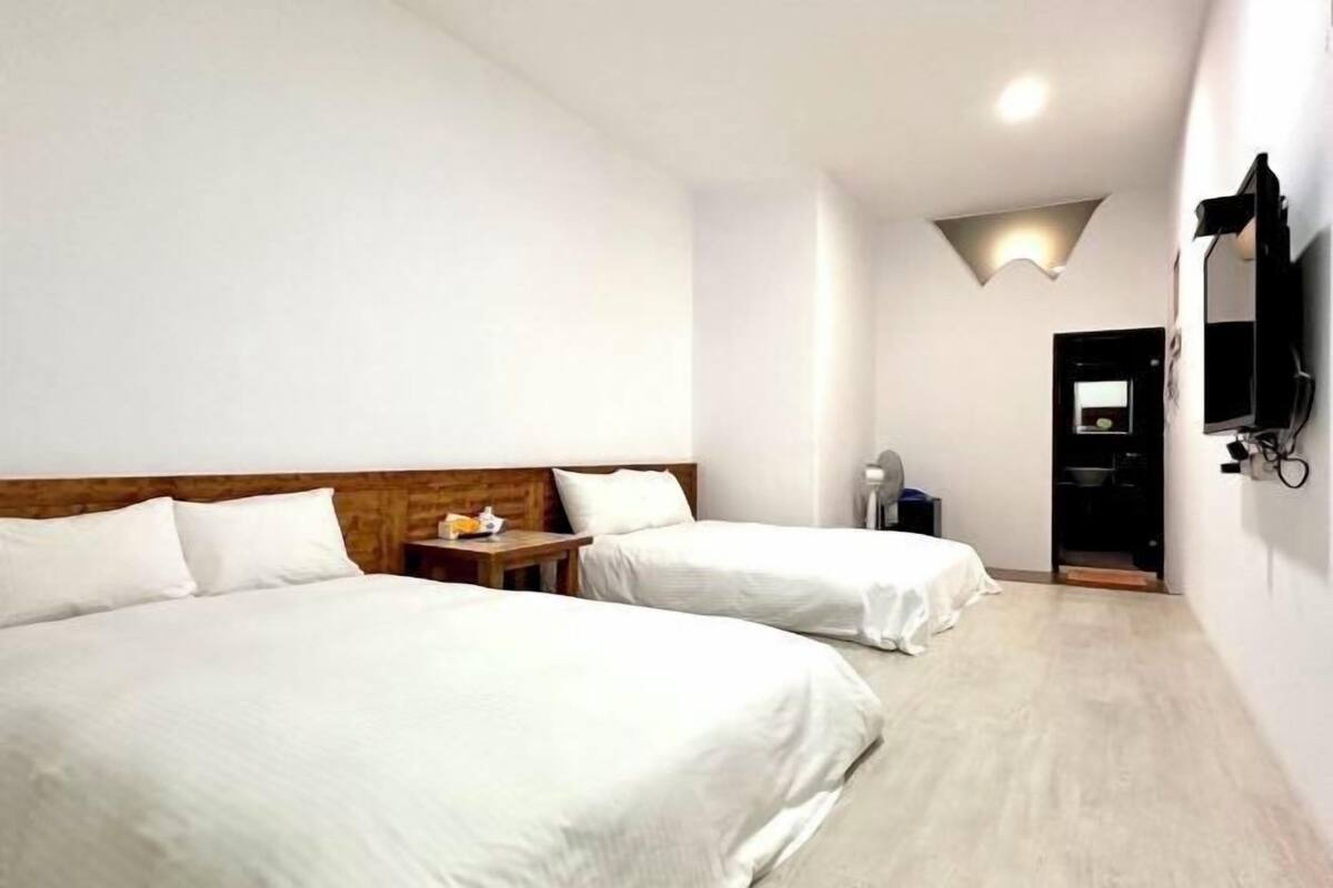 Una camera d'albergo con due letti, una testiera in legno, un tavolino con una lampada, una TV a muro e un ingresso che conduce al bagno.