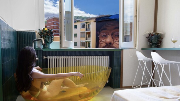Ett rum med ett stort badkar, en säng, ett fönster med utsikt över byggnader, ett element och ett litet bord med en vas.