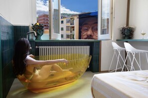 Quarto com uma banheira grande, uma cama, uma janela com vista para prédios, um radiador e uma pequena mesa com um vaso.