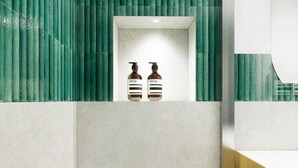 Ett badrum med en vägg täckt av gröna kakelplattor, en vit tvättställsinredning och två bruna flaskor med etiketten ”Aesop” stående på bänkskivan.