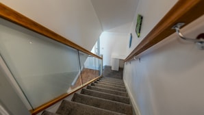 Una escalera con barandilla de madera y balaustradas de vidrio, que conduce a una habitación con paredes blancas y una puerta.