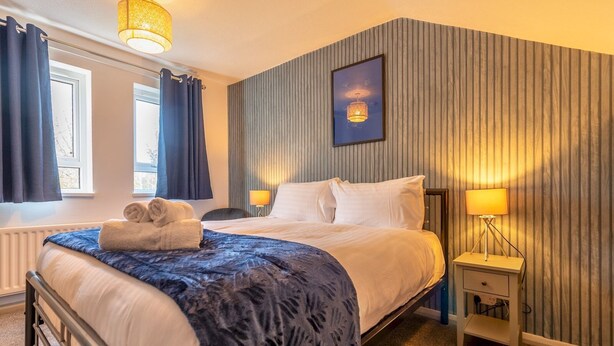 Una habitación de hotel con cama, dos lámparas de noche, una ventana con cortinas, un radiador y un cuadro en la pared.