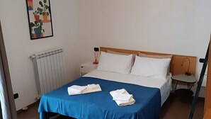 Un lit bien fait, avec une couvre-lit bleue et des oreillers blancs, une table de chevet avec une lampe et un radiateur au mur.