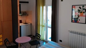 Un espace de vie compact comprenant un coin cuisine, un coin repas avec une table et des chaises, un radiateur et une fenêtre avec des rideaux.