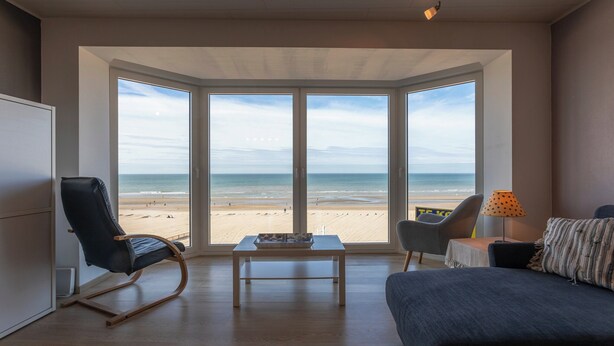 A room with a large bay window overlooking a beach, a bed, a desk, and a chair. - Cosy Renovated Flat on the Seafront (Koksijde)