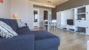 A modern living room with a dark blue sofa, a white dining table with chairs, and a white entertainment unit with a TV. - Cosy Renovated Flat on the Seafront (Koksijde)
