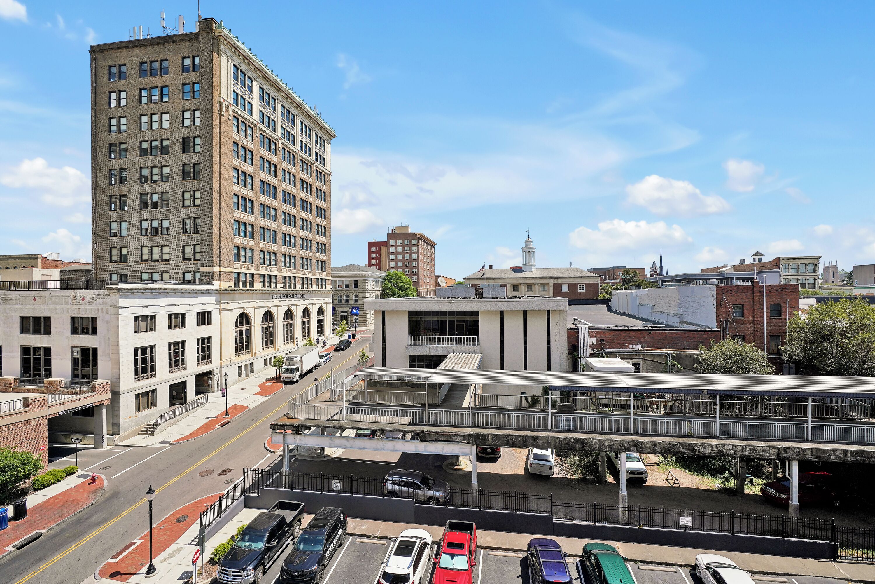 A multi-story building with a flat roof and large windows, adjacent to a street with parked cars and a pedestrian overpass.