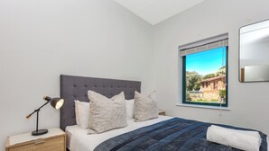 Ein Schlafzimmer mit einem Bett, einer Nachttischlampe, einem Nachttisch und einem Fenster mit Blick auf Gebäude.