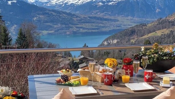 A table set with breakfast items, including fruits, bread, and drinks, overlooking a mountainous landscape with a lake.