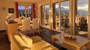 A modern living room with a sofa, a dining table with chairs, and a view of mountains through large windows.