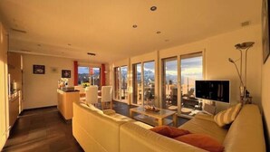 A modern living room with a sofa, a dining area, and large windows offering a view of the mountains.