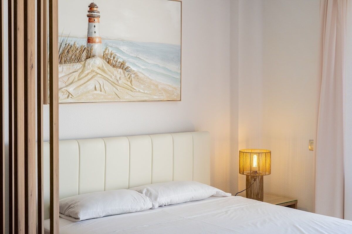 Una cama bien hecha con sábanas blancas, cabecero, lámpara de noche y un cuadro de un faro en la pared.