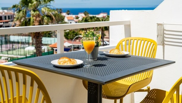 A balcony with a table set for breakfast, featuring croissants and orange juice, overlooking a coastal view. - Modern studio with ocean views and heated pool in Malibu Park (San Eugenio Alto)